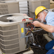 HVAC technician in yellow hard hat repairs outdoor AC unit, inspecting wires. Multiple air conditioning units on site in British Columbia.
