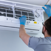 An HVAC technician in blue gloves cleans an air conditioner, lifting the panel to access filters in a British Columbia home.