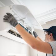 A technician in gloves and a cap checks a wall-mounted ductless mini-split AC, lifting the panel for HVAC service in British Columbia.