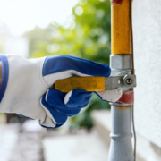 A gloved hand adjusts a yellow gas line valve outside a home in BC, near a white wall and green plants—HVAC service in action.