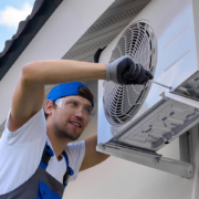HVAC technician in safety gear repairs an outdoor heat pump unit mounted on a building wall in British Columbia.