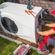 HVAC technician in red uniform services outdoor heat pump beside a brick wall and grass, showcasing expert BC heat pump repair.