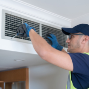 An HVAC technician in safety gear services an indoor air vent with a screwdriver to improve air quality in a British Columbia home.