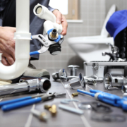 A plumber in a bathroom uses a wrench on a white pipe under the sink, surrounded by plumbing tools—HVAC service in BC.
