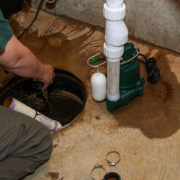 HVAC technician in a British Columbia basement repairs a sump pump, hand in pit, with pipes, water, and tools on the concrete floor.