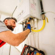 HVAC technician in British Columbia repairs a wall-mounted gas boiler with tools, pipes, and wires visible in the background.