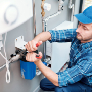 Plumber in blue cap and plaid shirt using a wrench to service a water filter, pipes and outlets visible—HVAC in British Columbia.