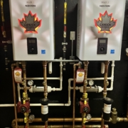 Two CANUCK Navien boilers with maple leaf logos are installed side by side on a black wall, featuring copper pipes and HVAC gauges.
