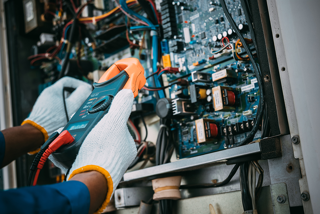 An HVAC technician in white gloves uses a clamp meter to inspect wiring and components inside an open control panel in BC.
