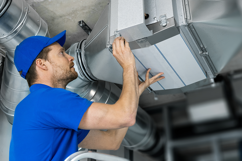 HVAC technician in blue uniform checks and repairs ceiling air duct with tools in a commercial building in British Columbia.