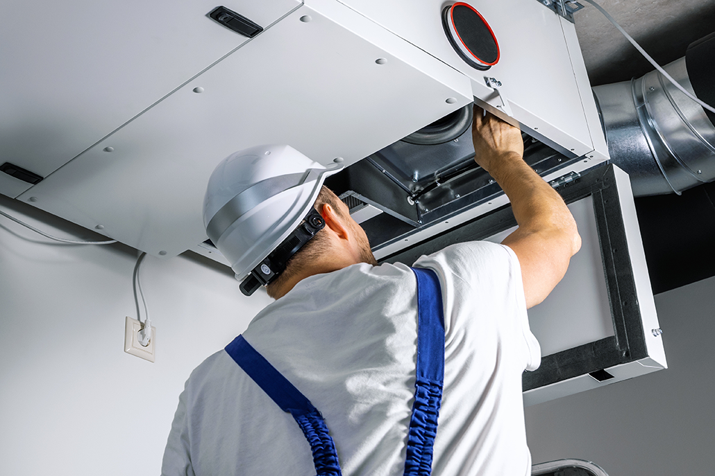 HVAC technician in a white hard hat and blue overalls repairs a ceiling-mounted HVAC unit with tools in British Columbia.