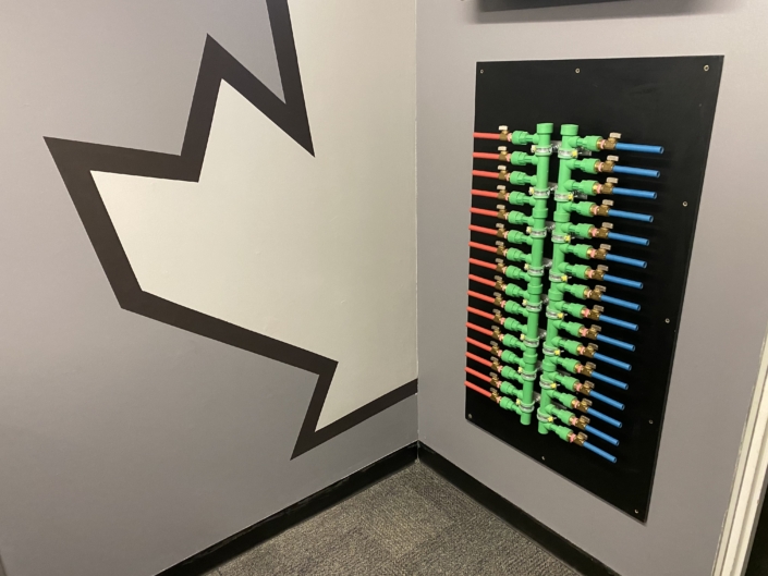 Wall-mounted HVAC panel with green pipes, color-coded tubing, and brass valves in a gray BC room, abstract art on wall.