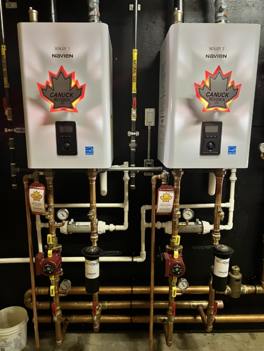 Two Navien boilers by Canuck Plumbing, side by side on a black wall with copper piping, in a BC HVAC service setup.