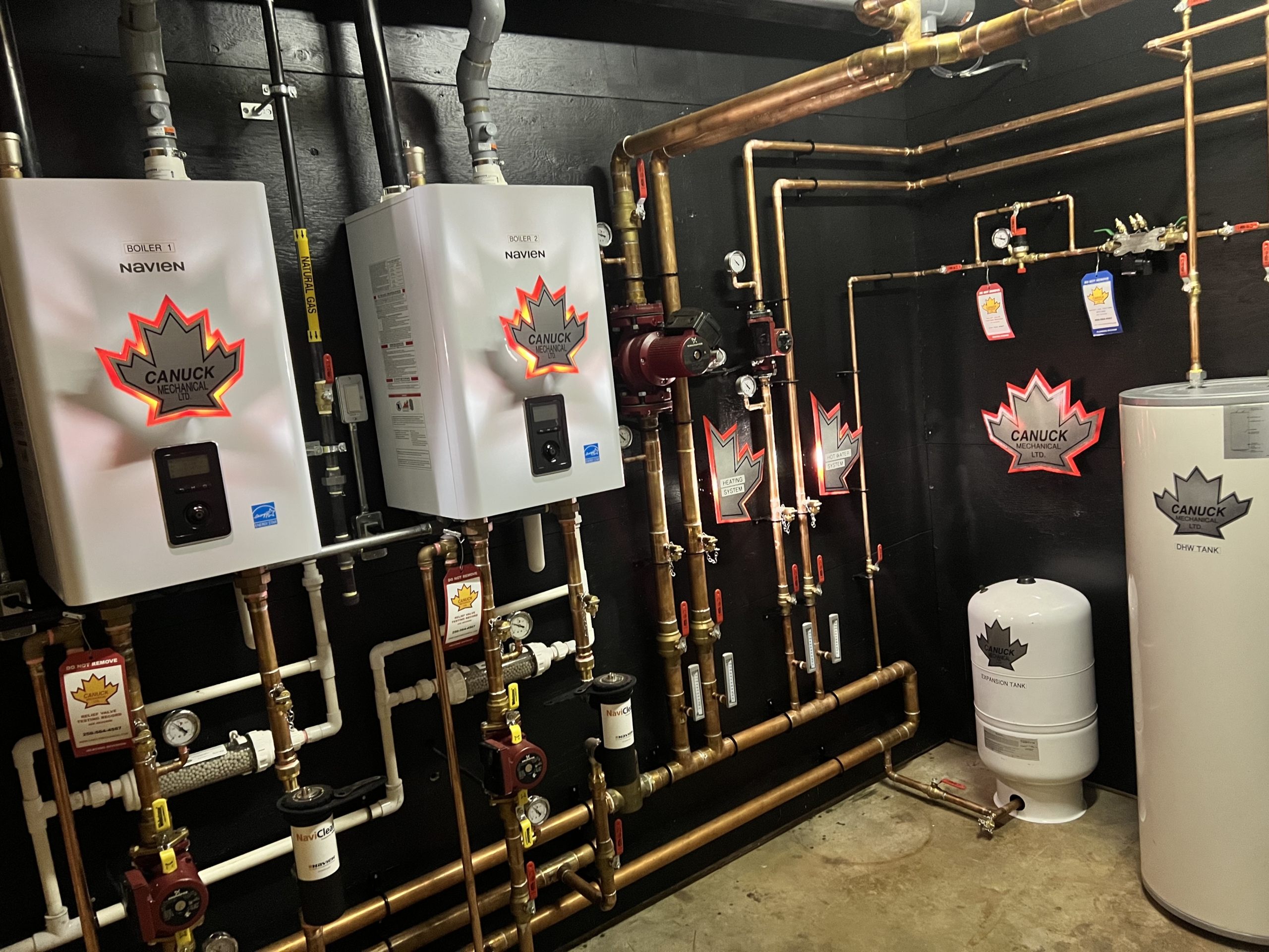 Mechanical room with two wall-mounted Navien boilers, copper piping, expansion tank, gauges, and Canuck Mechanical logos—BC HVAC experts.