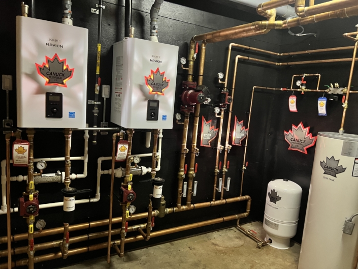 Mechanical room in BC with two Navien water heaters, copper piping, a white tank, valves, and Canuck Plumbing maple leaf stickers.