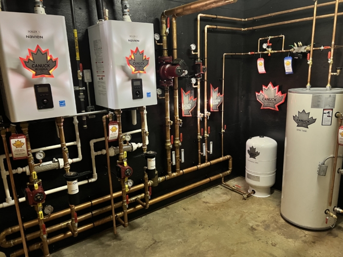 British Columbia HVAC room with two Navien water heaters, water tank, pipes, gauges, labeled parts on black walls and Canuck decals.