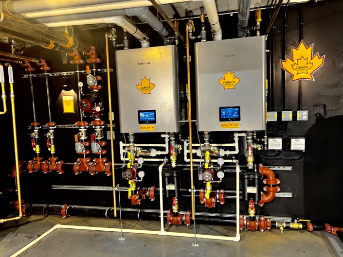 Boiler control panels with digital screens and Canuck branding, surrounded by pipes and valves, in a BC HVAC system setup.